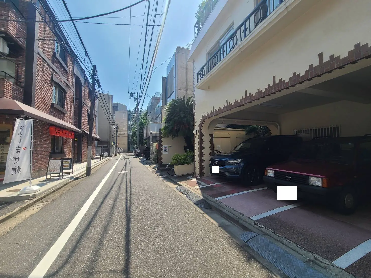 - 日差しが降り注ぐ静かな街並み、レンガ造りの建物と駐車場に停まった車がある風景