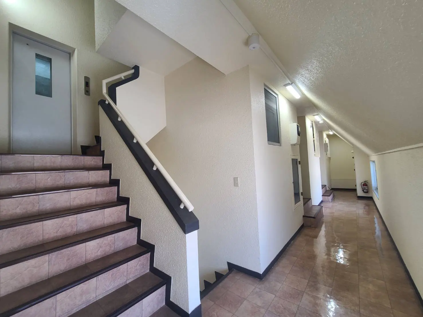 - シンプルで明るいデザインの階段、白い壁とタイルの床、手すり付きの階段とエレベーターの入り口がある廊下の様子