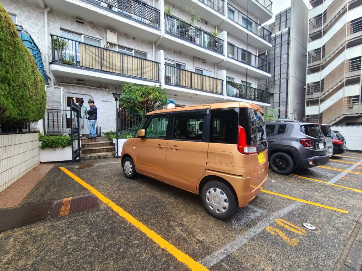 駐車場に停まっている茶色の箱型車両と周囲の複数の車、背景にはアパートメントの建物と歩道に立つ人物
