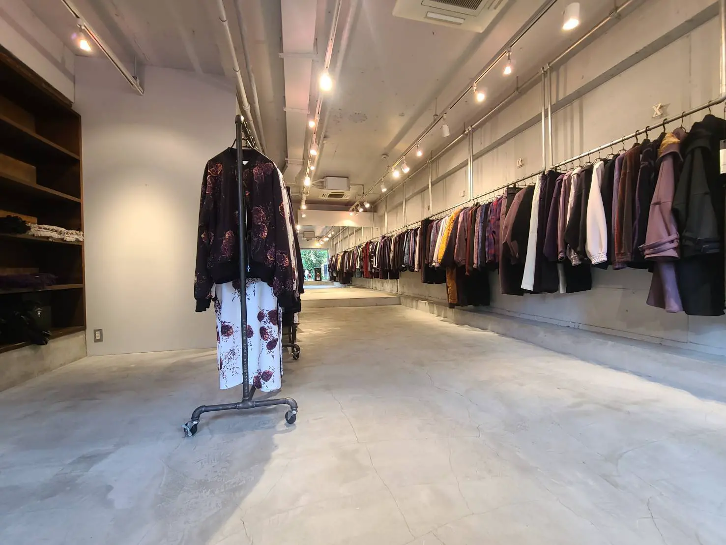 シックなデザインの洋服店内、コンクリートフロアにユニークな照明、壁に並ぶ様々なカラーのアパレル商品