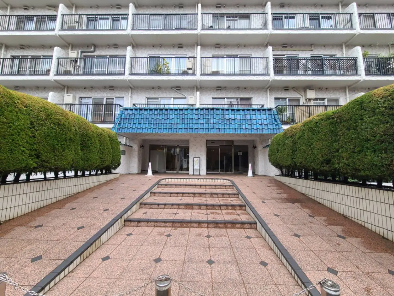 白い壁の外観を持つマンションのエントランス、整備された緑の生垣と青い屋根が特徴的