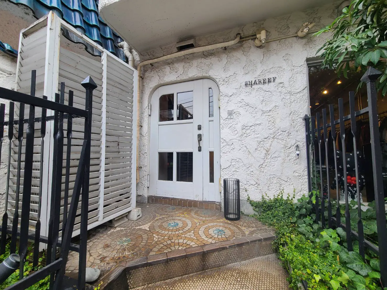 モザイクタイルが敷かれたエントランスと白い壁の外観、緑植物が配されたおしゃれな店舗入口