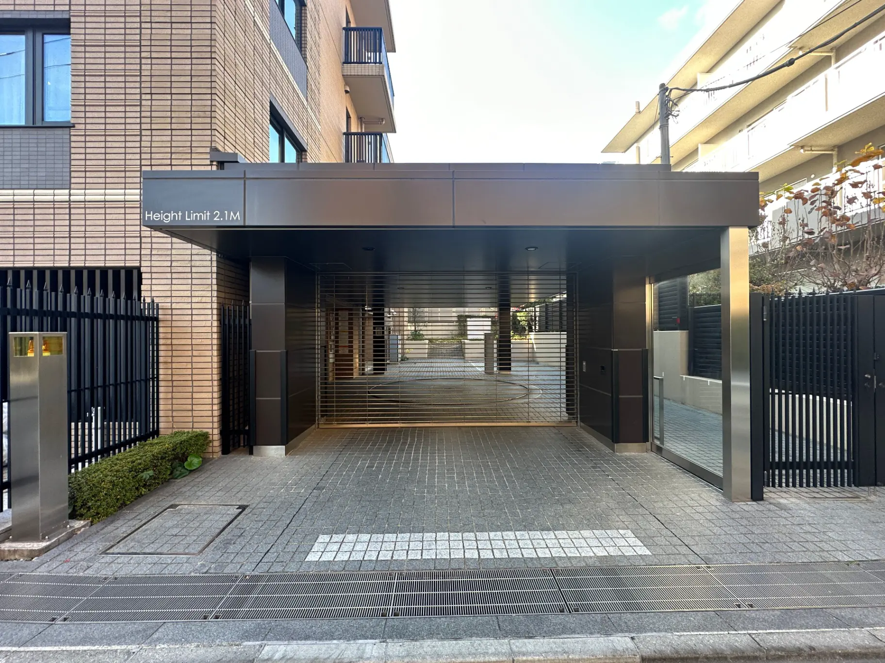 現代的な外観の建物入口、車高制限2.1メートルの標示付き、黒い門扉とタイル張りの歩道