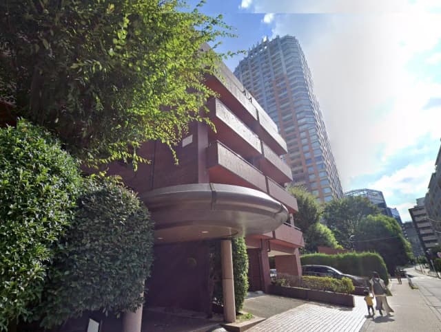 都市部に位置するモダンなマンションのエントランス、周囲の緑と歩行者、晴れた日の青空
