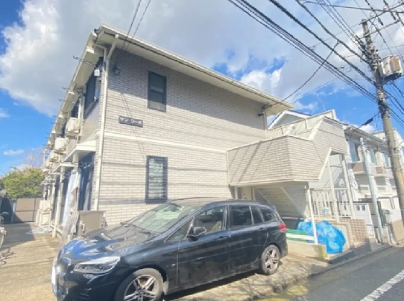 - ブロック状の外壁を持つアパートメントの外観、前には黒い車が停まっている、青空と白い雲が広がる背景