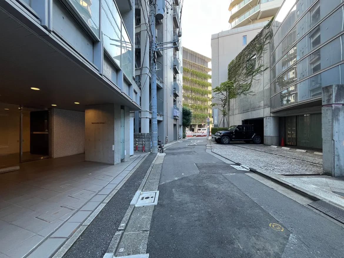 - コンクリートの建物に囲まれた静かな通り、左側にガラス張りのエントランスと車が停まる駐車場の様子