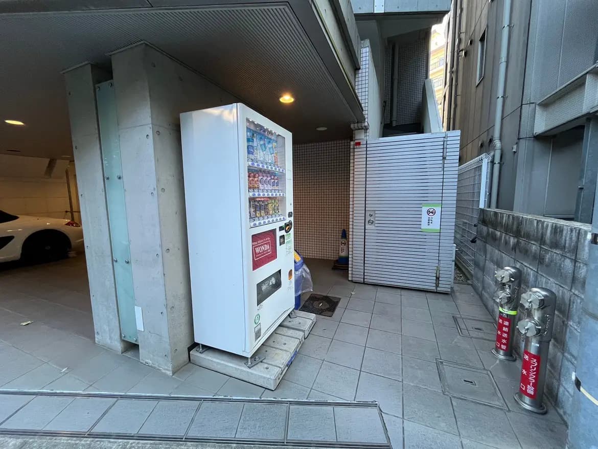 - 駐車場の出入り口付近にある自動販売機、背後には白い車とストレージ用の扉が見える屋外のシーン