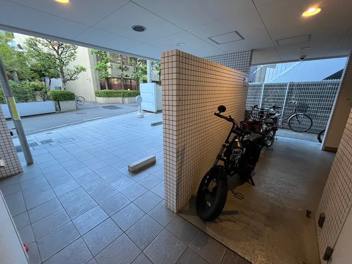 - 駐輪場の入り口、モダンなタイル張りの床に並ぶ自転車と電動バイク、周囲に緑が見える風景