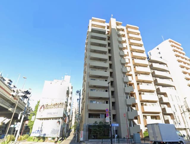 - 高層マンションの外観、青空を背景にした建物と近隣の店舗が見える街並み