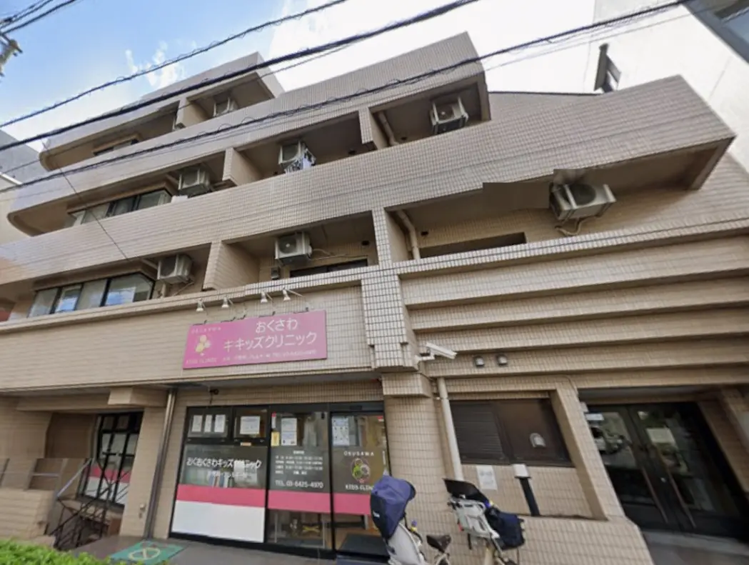 - おくさわキッズクリニックの外観、明るい色合いの看板や空調設備が特徴の病院ビル、周囲には自然が広がるエリア