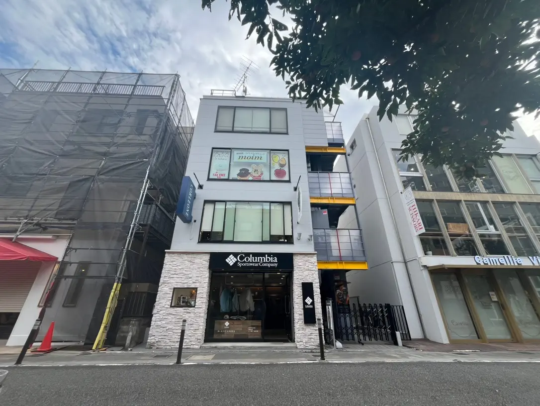 都会の通りに面した現代的なビル群、アパレルショップのColumbiaブランドの旗を掲げる店舗、曇天の下の日常風景