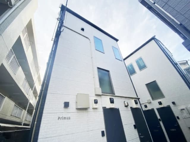 - 白い外壁の建物と隣接するアパート、明るい空の下での外観、Primusの名称が見えるエントランス。