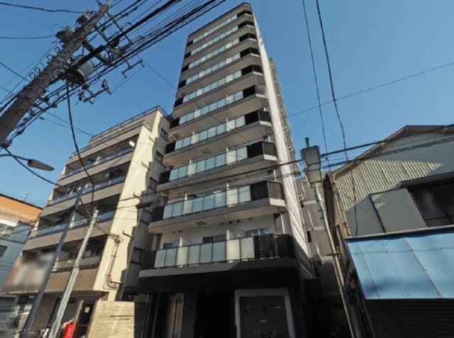 - モダンなデザインのマンション外観、ガラスバルコニーと青空、周辺には電柱と商業施設が見える景観