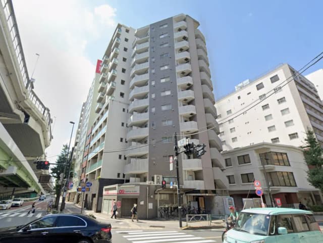 - 都市部に建つ現代的なマンション、周囲には交通信号や商業施設、歩行者の姿が見える街の風景