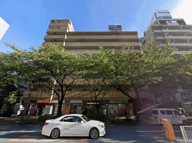 - 緑豊かな街並みに佇む複数階建ての商業ビル、白い車が通る道路、晴れた青空の下で撮影された景観