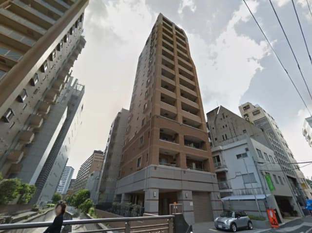 - 高層マンションの外観、周囲に並ぶビル群と青空、手前には歩道と車両が見える都市の風景