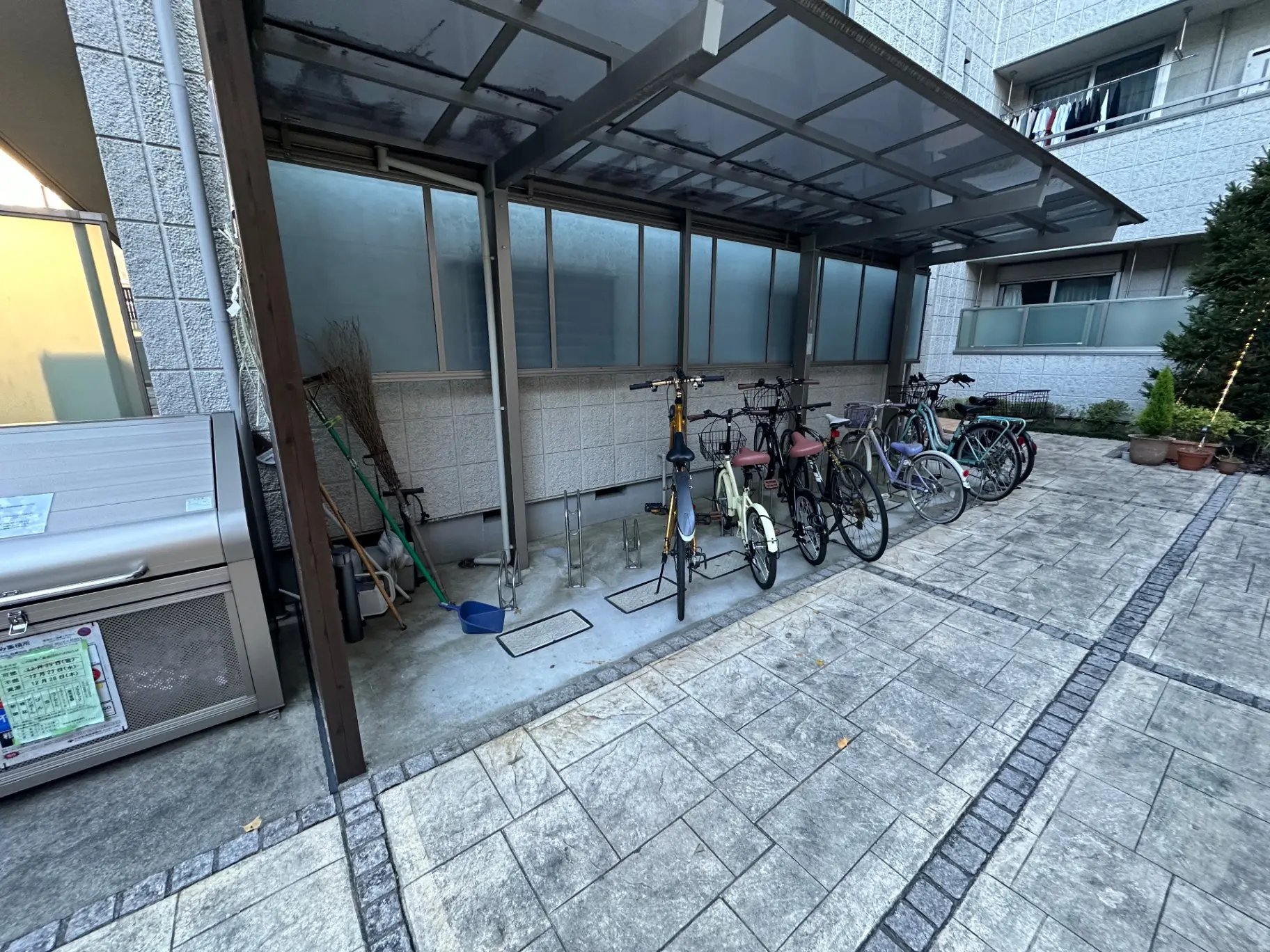 アパートの屋外駐輪場、整理された自転車、屋根付きスペース、背景には建物と植木