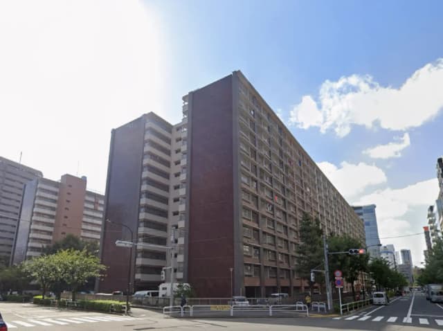 - 高層住宅の外観、青空と雲が広がる中、周囲に緑の木々と交通標識が並ぶ街の風景