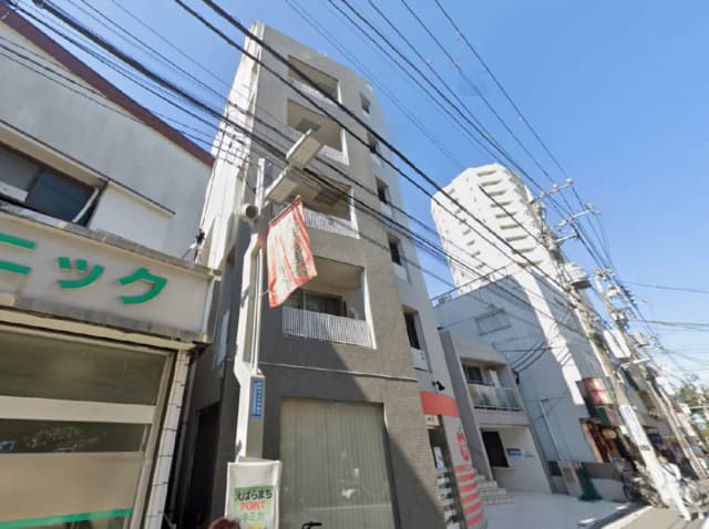 - ビルの外観、電線が張り巡らされた街並みと青空、近隣の店舗が見える立地