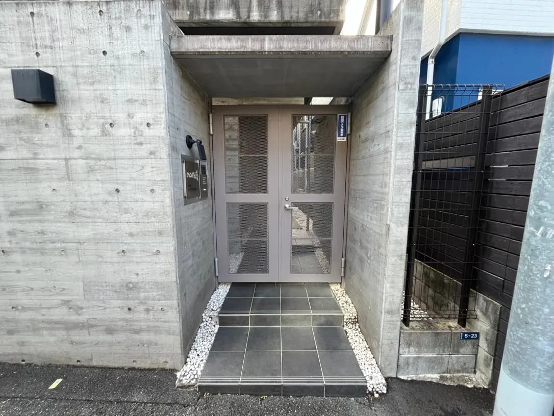 コンクリート打ち放しのデザインが特徴的なエントランス、黒いタイルの地面とモダンな灰色のドア