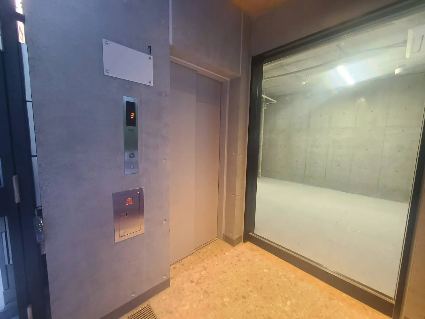 - コンクリート打ちっぱなしのエレベーター前、階数表示パネルとスイッチ、ガラス窓越しに見える無機質な空間の様子
