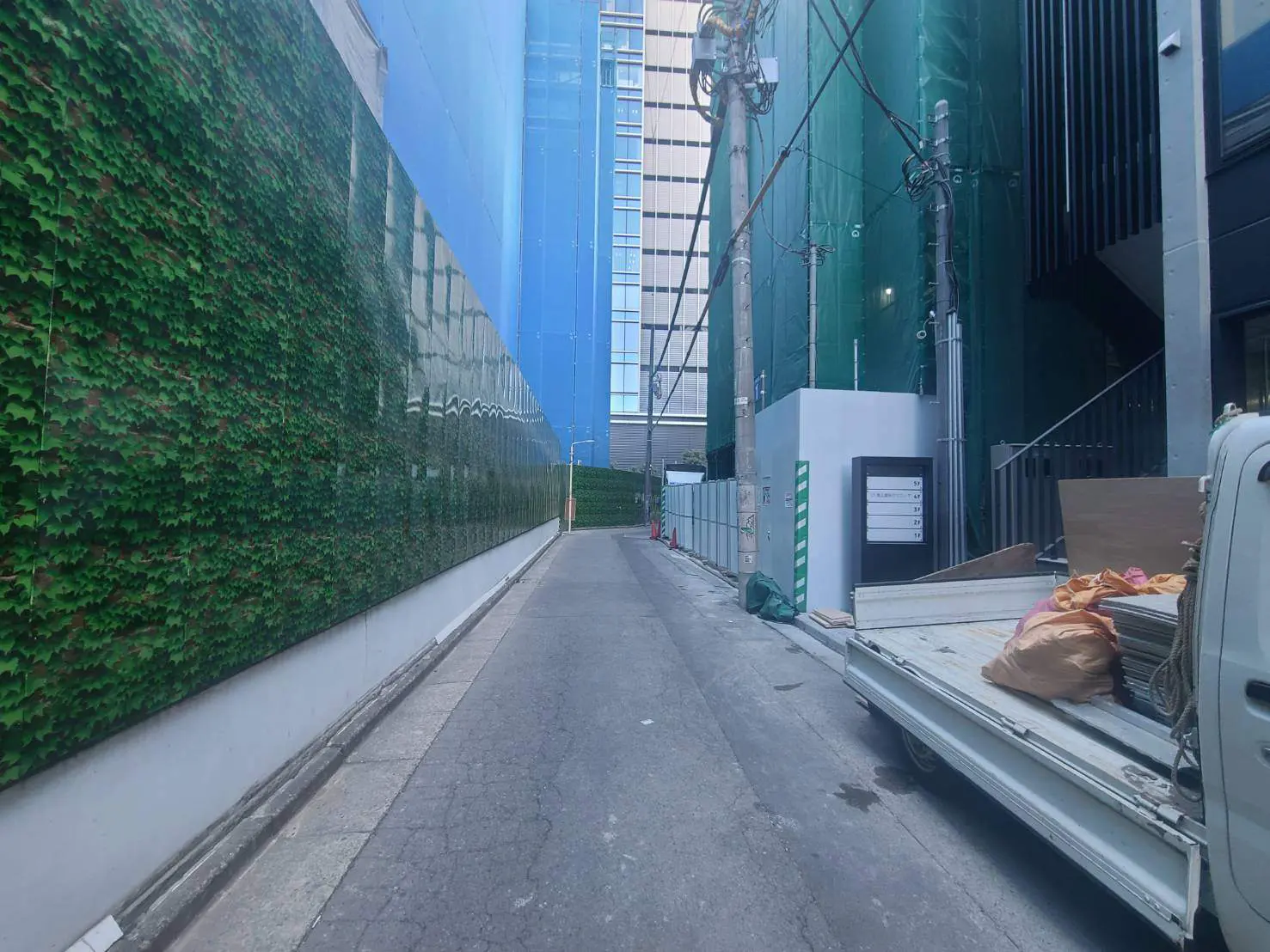 - 緑の壁面を持つ狭い路地、建設現場の背景と白いトラックが並ぶ都市の風景