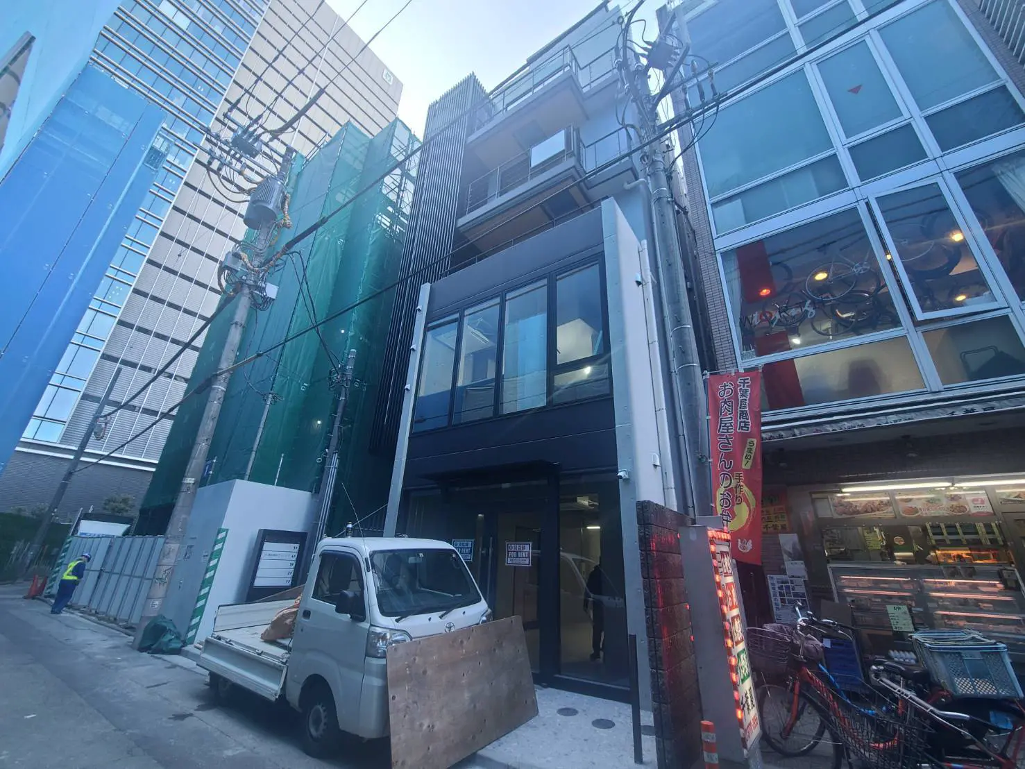- 新しいビルと工事中の建物が並ぶ都市の風景、手前には白い軽トラックと飲食店の看板が見える通りの様子