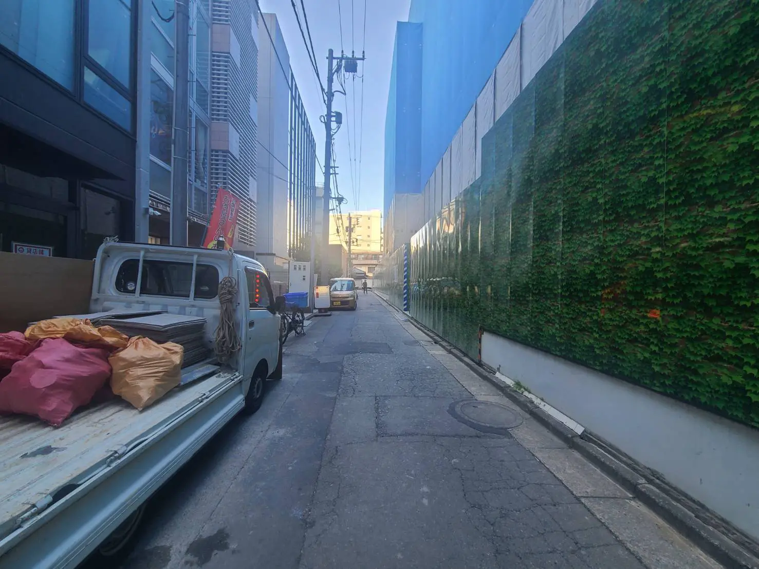 - 板材と荷物が積まれたトラックが停車している街の狭い路地、背景には商業ビルと緑のフェンスが見える風景