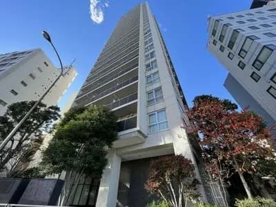 - 高層マンションの外観、周囲には緑豊かな植栽と都市的な建物が立ち並ぶ晴れた日中の風景