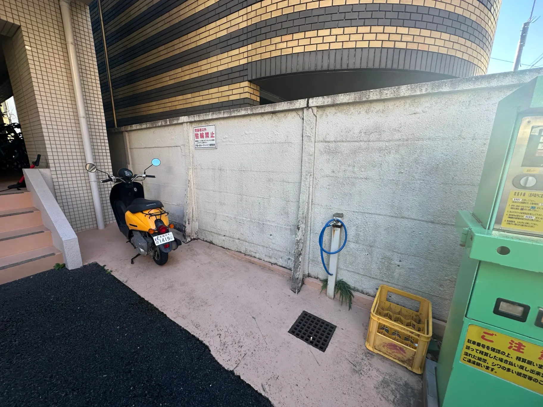都市部の駐輪場、黄色いスクーターが停められている様子、コンクリート壁と緑のホースのある景色