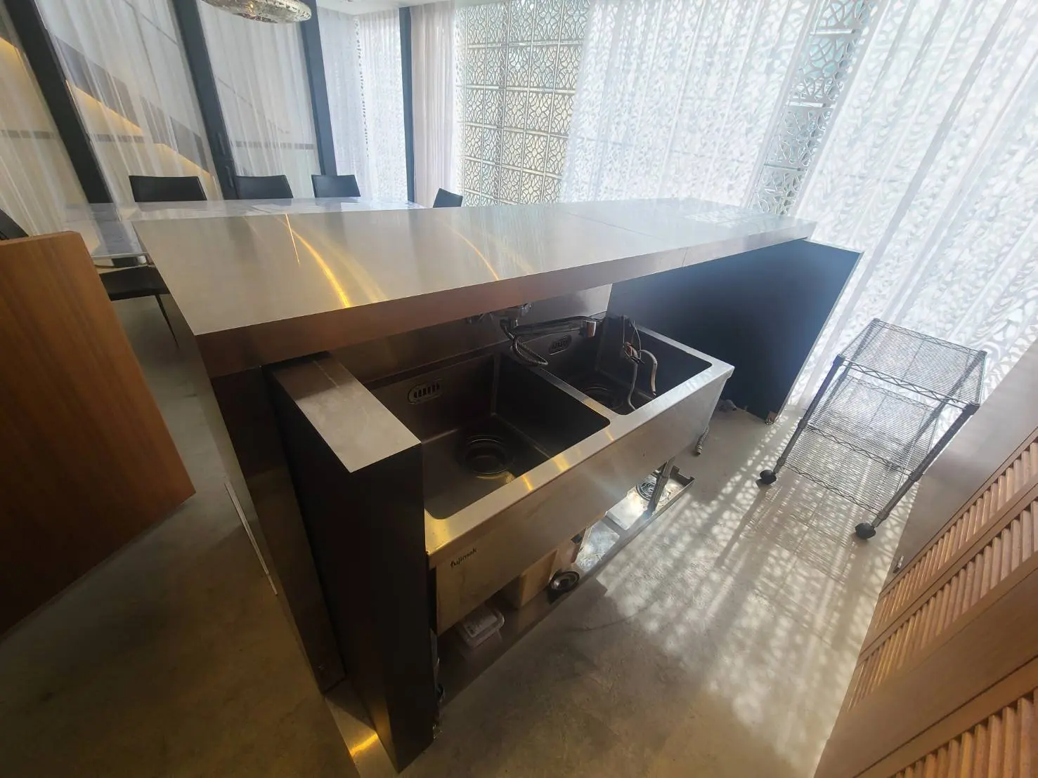 光が差し込むモダンなキッチン、ステンレスの作業台とシンク、繊細な模様のレースカーテン