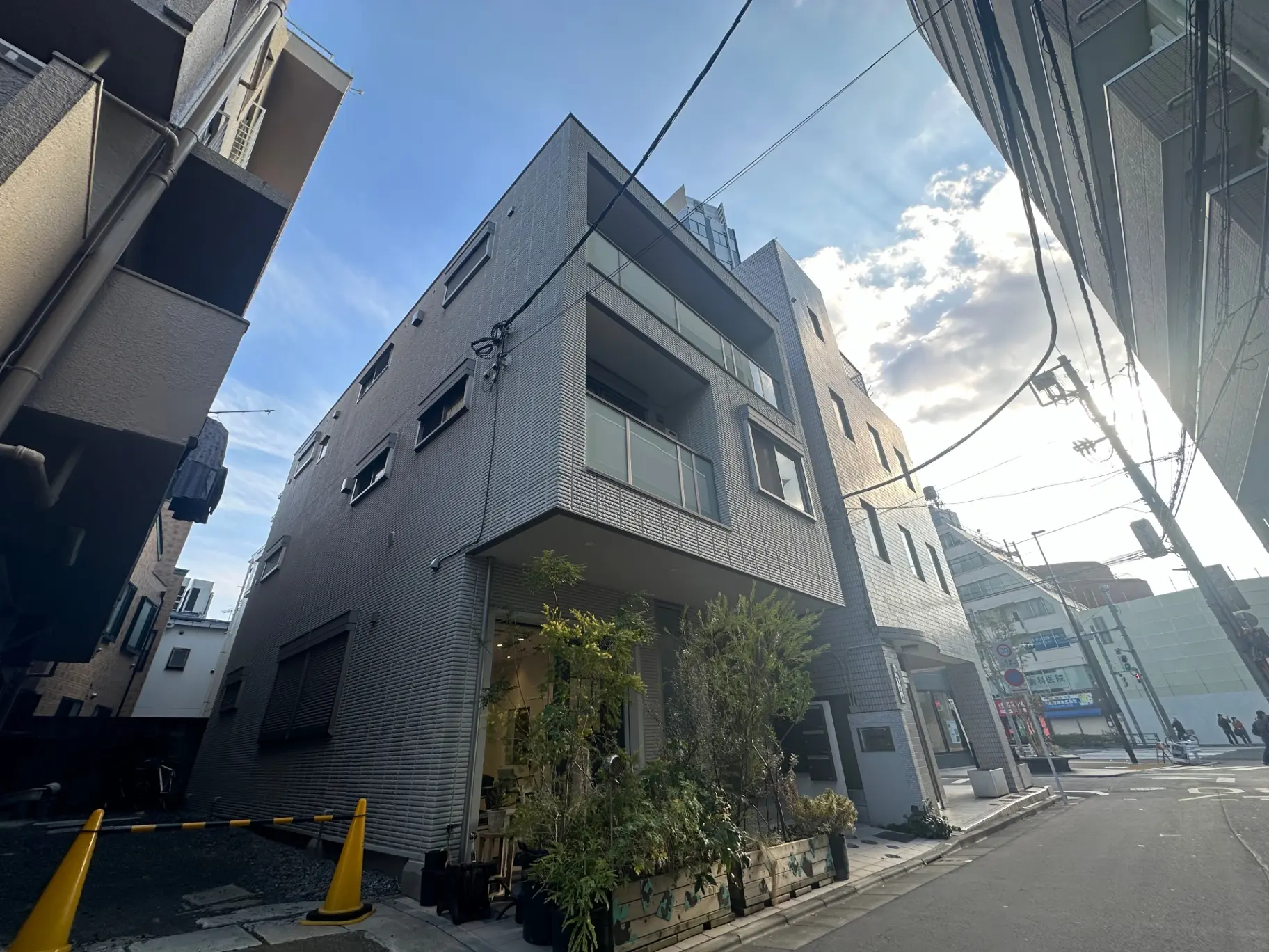 都会的なモダンな外観の建物、グレーの壁とガラスの窓、一階には植物が飾られたおしゃれなカフェ