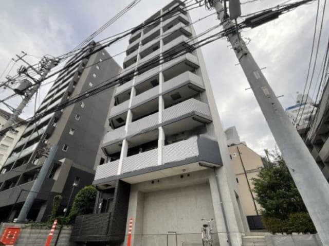 - モダンな外観のマンション、白とグレーのバルコニー、草木のあるエントランス、周囲には電線が走る都市景観