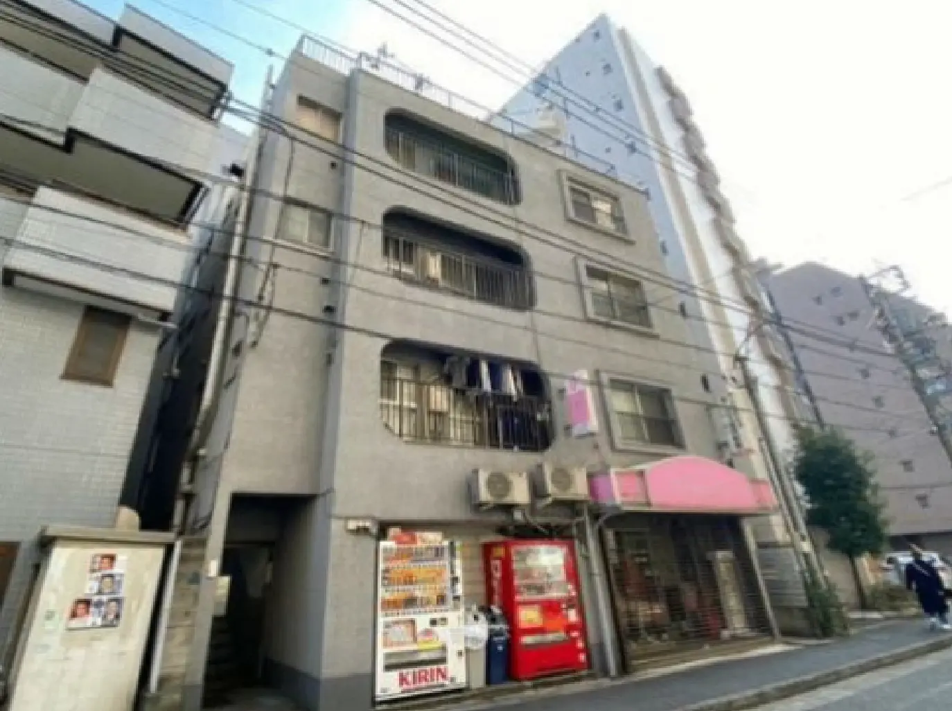 - 薄いグレーの外壁を持つ古いアパート、下には自動販売機と店舗の看板、周囲には高層ビルが立ち並ぶ風景