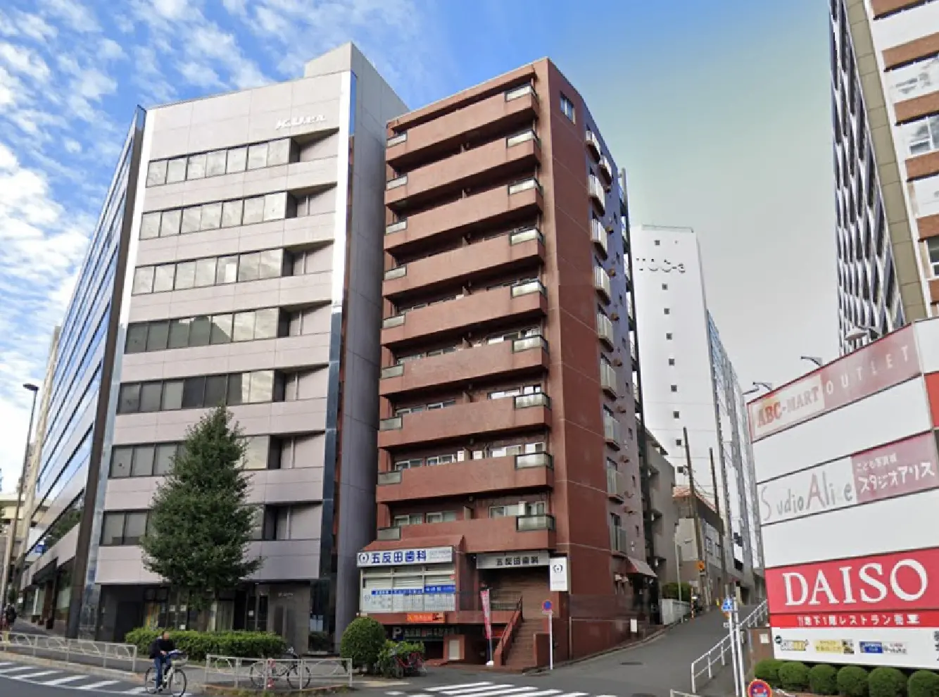 - 近代的なビルが立ち並ぶ街並み、赤い外壁のマンション、周囲には緑の木と自転車を使う人々の風景