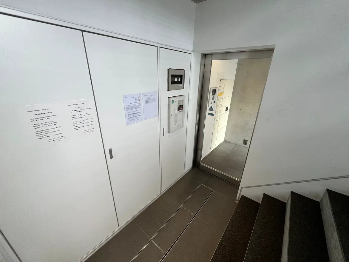 - ホワイトの収納ドアとエレベーターの扉があるシンプルなエントランスホール、壁には注意事項の掲示物が貼られている