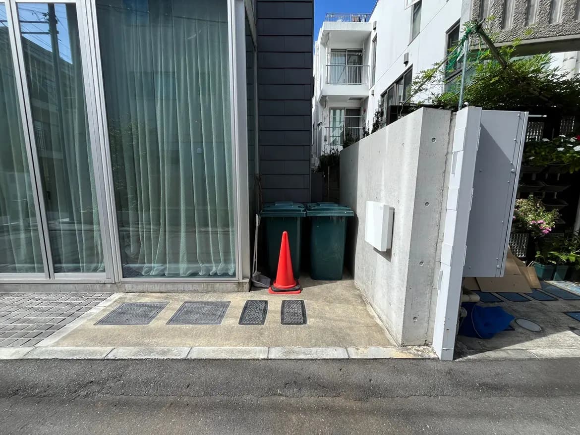 - 厳選されたゴミ箱と赤いコーンが並ぶ狭い通路、透明なカーテンがかかるガラス壁、近隣の住宅が見えるシティライフの風景