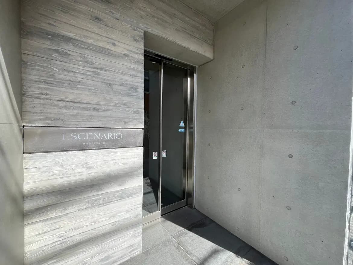 - モダンな外観の建物入り口、ナチュラルウッドの仕上げとコンクリートの壁、エレベーターのドアが見えるシーン