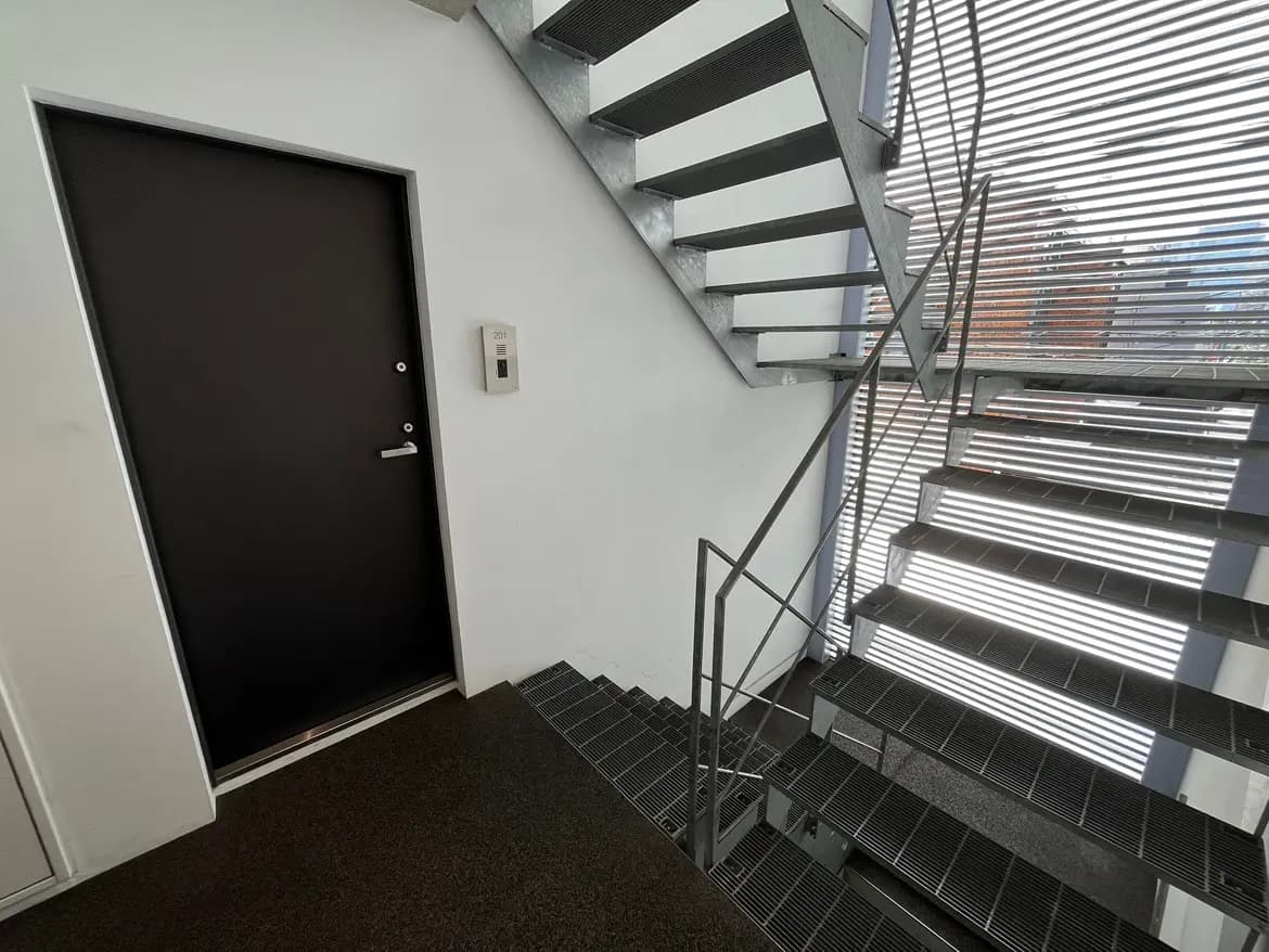 - モダンな建物の階段、ガラスの手すり、黒いドアとインターホンがあるシンプルなエントランスの風景