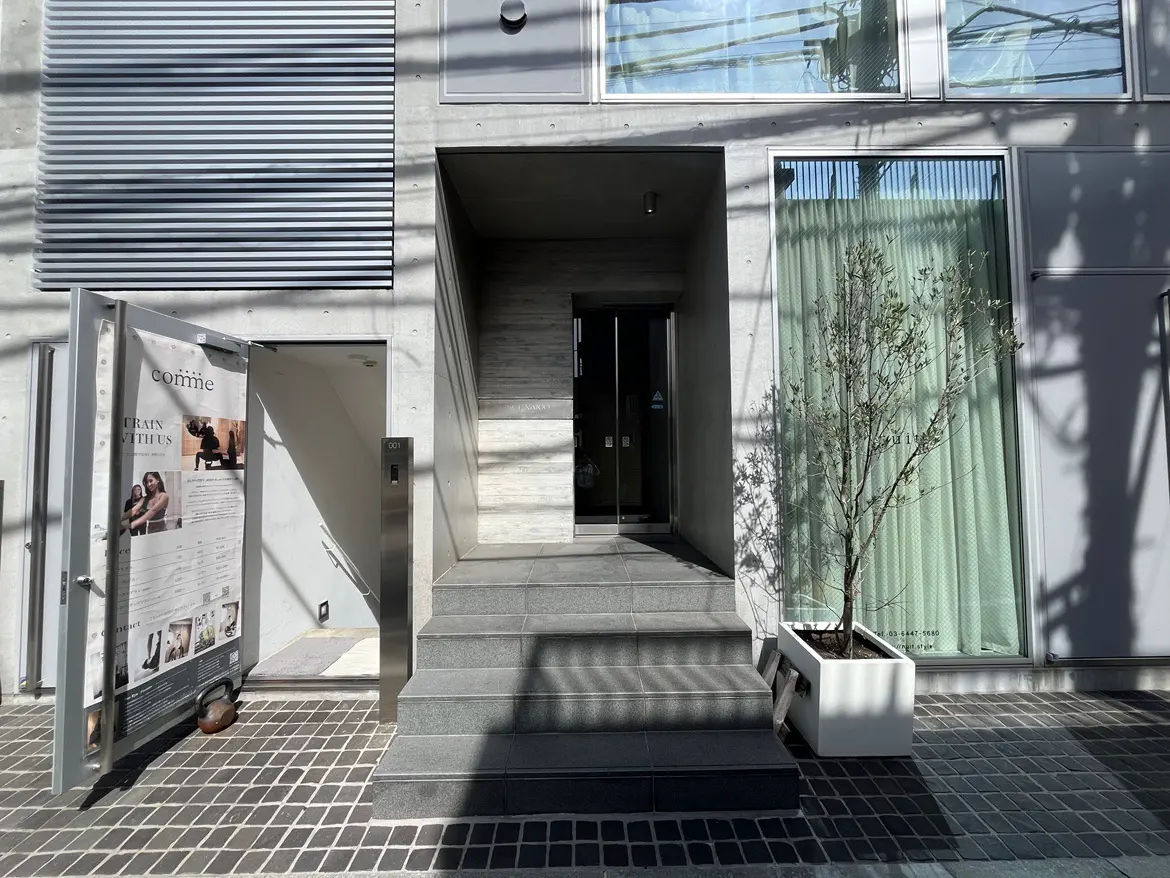 - モダンなエントランス、スロープ付きの階段、開いたドアと案内板、オリーブの木が置かれたスタイリッシュな外観