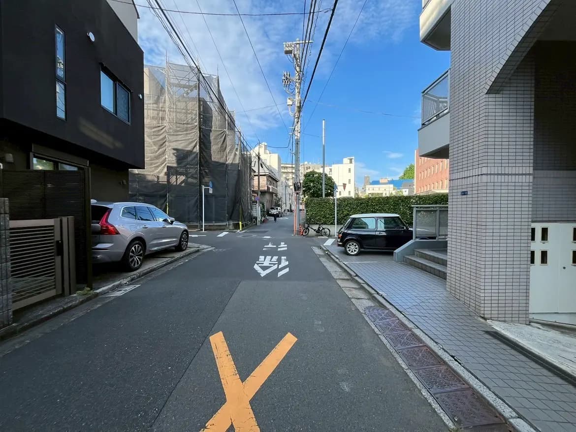 - 都市の住宅街の一角、青空の下に広がる舗装された道路、両側に並ぶ建物と停車した車両が見える風景