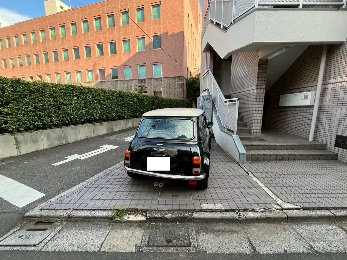 - 黒いミニカーが歩道に駐車されている様子、背後には植え込みとビルの壁が見える街の風景