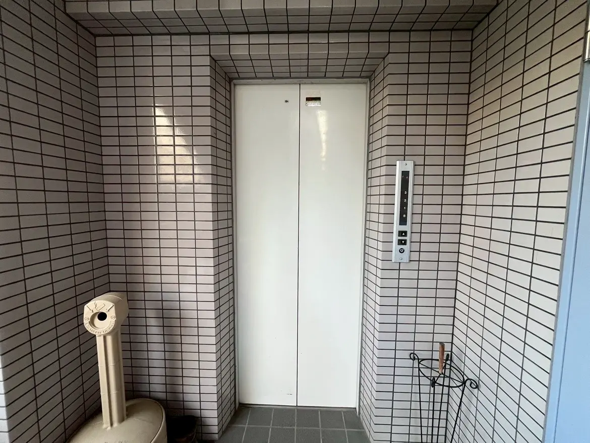 - タイル張りのエレベーター前、白いドアとエレベーター操作パネル、ゴミ入れと傘立てがあるエントランスのインテリア