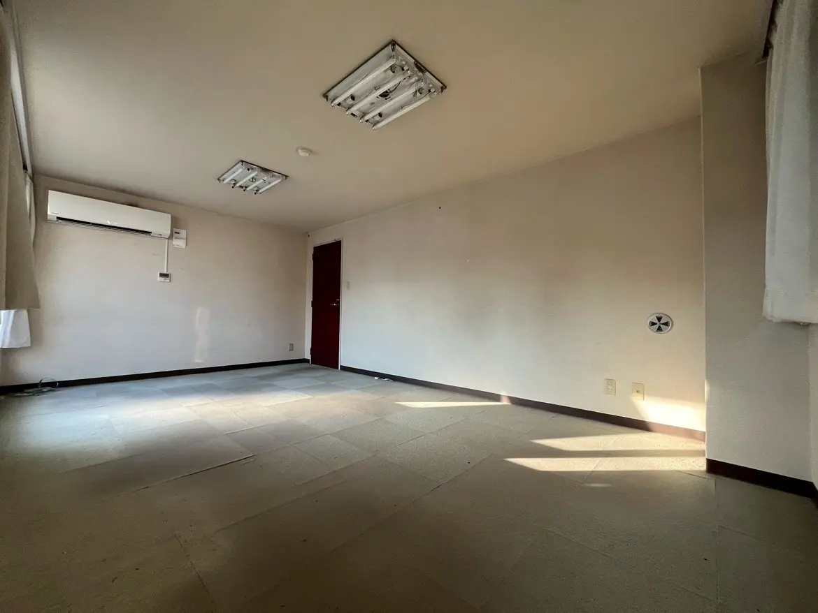 - シンプルなデザインの空き部屋、白い壁と窓からの自然光、エアコンとドアがある室内の様子