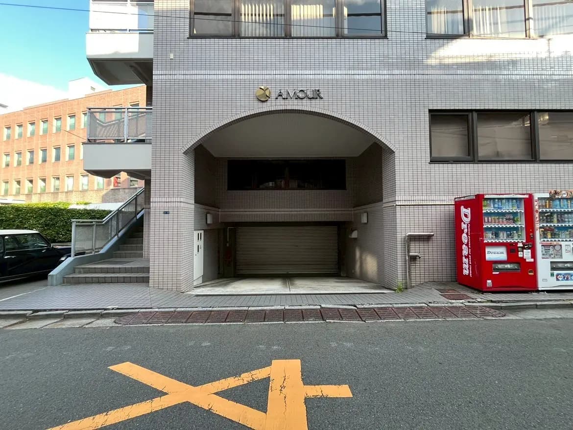 - ブロックタイルで外装されたアパートのエントランス、看板「AMOUR」、自動販売機と傾斜のある階段が見える通りの風景