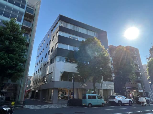 - 明るい青空の下に立つ現代的なビル、シンプルなデザインの外観と周囲にある緑豊かな木々の風景