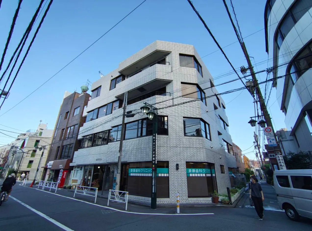 - 白いタイルを使用したモダンな外観のビル、近くには飲食店や医院があり、青空の下で人々が通行する街並み