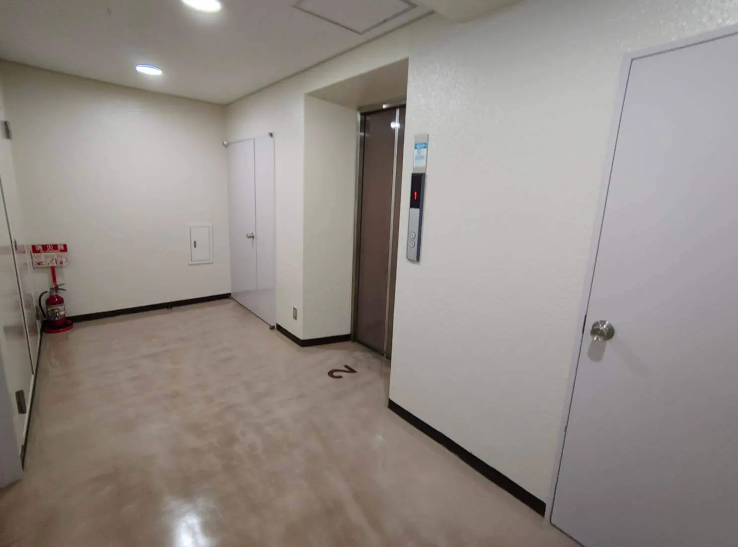 - エレベーターと白い壁、階数表示がある廊下の内部、中間に防火器具が設置されたシンプルなインテリア