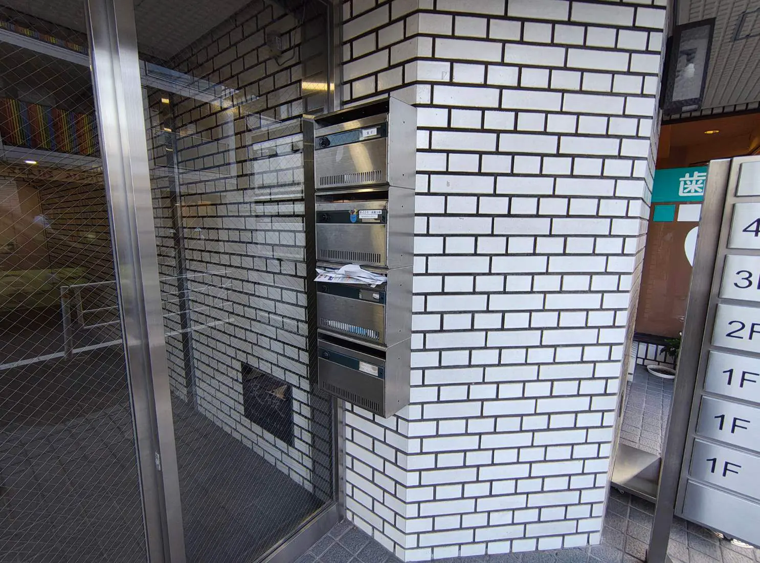 - 白いタイル張りの壁に取り付けられた郵便受け、周囲はガラスのドアで囲まれたエントランスの様子