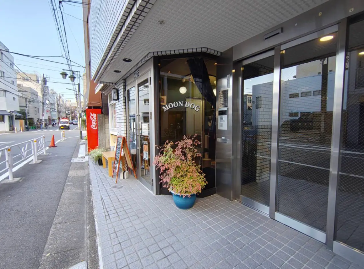 - 「MOON DOG」の入り口、グレーのタイルと植え込み、ガラス扉の外観、近くの通りと飲食店の風景